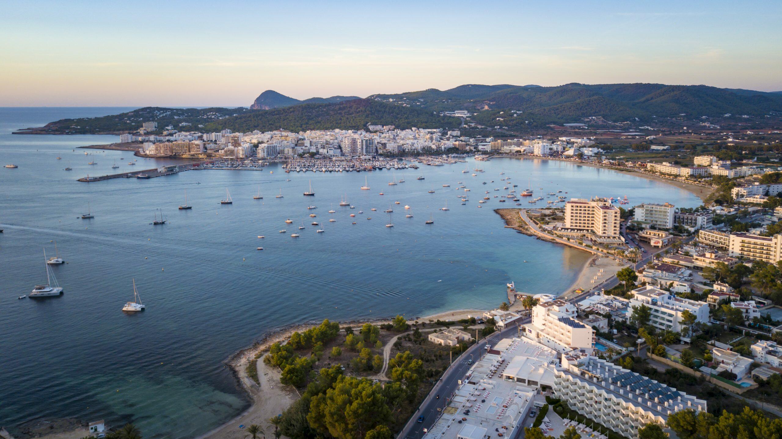 DJI_0095 Aerial view of San Antonio Bay, Ibiza, with anchored sailboats, a marina and waterfront buildings backed by wooded hills.